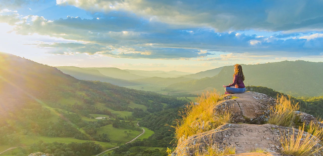 Woman meditating on top of a rock at the mountains in Brazil with a beautiful sunset, blue skies with clouds and the mountains in the horizon